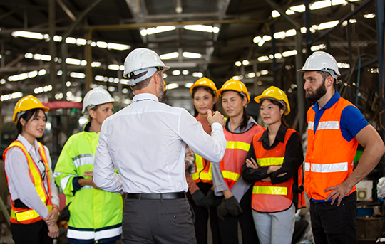Safety officer briefing a group of workers wearing helmets and safety vests at a construction site.